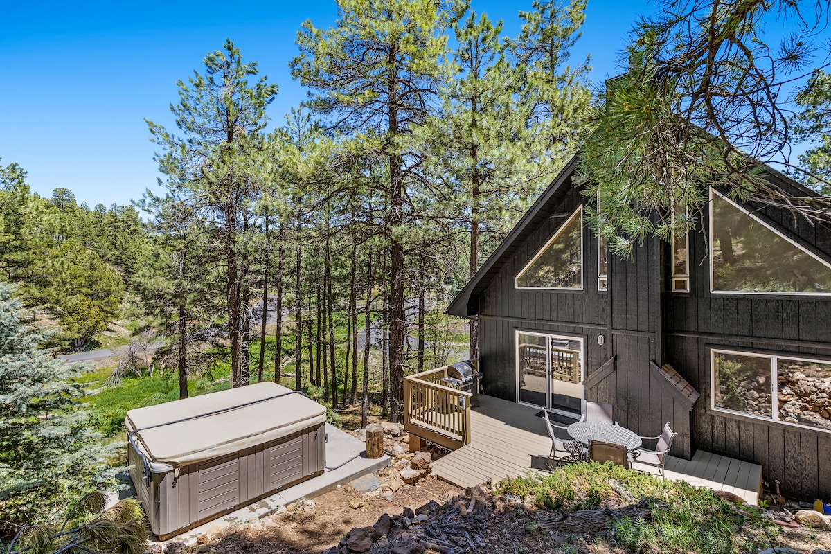 The exterior of the cabin is visible, featuring a spacious wooden deck that leads to a hot tub. Tall pine trees surround the property, while the cabin's large windows reflect the natural light. A pathway indicates proximity to the nearby landscape.