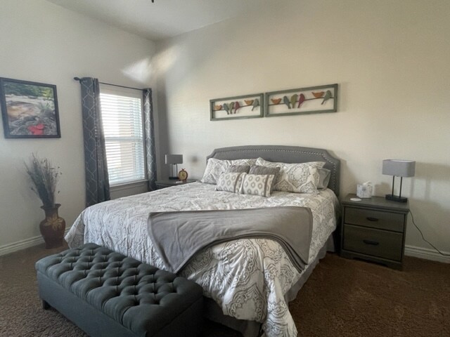 The spacious master bedroom features a king-sized bed with decorative pillows and a cozy gray blanket. A tufted bench is positioned at the foot of the bed. Nightstands with lamps flank the bed, and natural light filters through the window with sheer curtains.