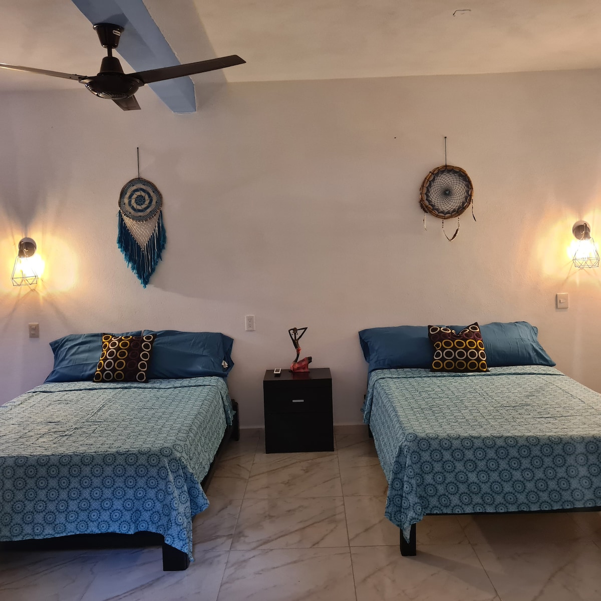 Two spacious beds are positioned against a neutral wall, each adorned with blue bedding and decorative pillows. Ambient lighting is provided by wall-mounted lanterns, while decorative wall hangings add a touch of character to the room. A small bedside table is located between the beds.
