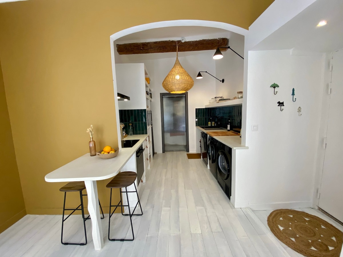 A modern kitchen is presented with a warm golden wall and whitewashed wooden flooring. A bar with two stools is located on the left, while appliances and ample storage can be seen along the wall. Decorative lighting fixtures add a subtle enhancement to the space.