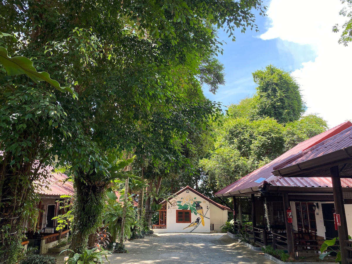 A pathway is visible, framed by lush greenery and tall trees. On either side, distinct buildings with red roofs are situated among the foliage, contributing to a tranquil atmosphere. The sky above is clear and bright, enhancing the natural beauty of the surroundings.