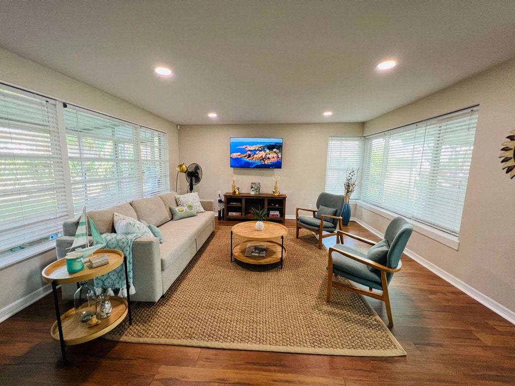 A spacious living area is presented with natural light streaming through multiple windows. A comfortable sofa faces a mounted smart TV. A round coffee table sits at the center, surrounded by armchairs. A decorative fan and a few plants add to the welcoming ambiance.
