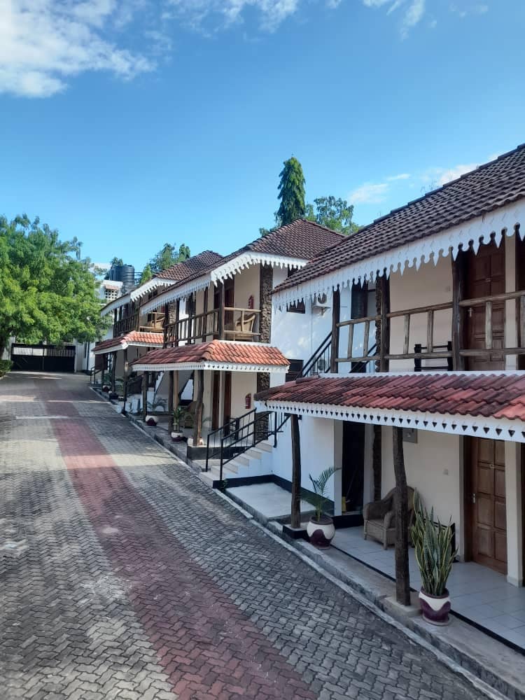 A row of charming guest rooms is presented with traditional architecture. Each room features a private balcony with wooden railings. Paved pathways and lush greenery create a serene outdoor environment, complemented by clear blue skies above.