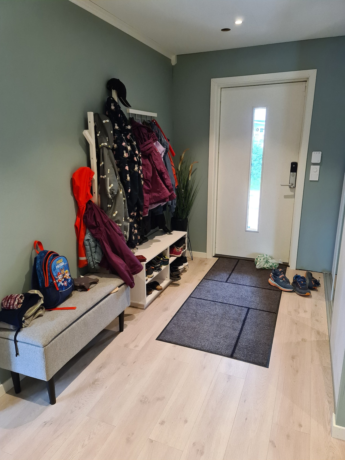 A functional entryway features a light-colored bench with a shoe rack and a wall-mounted coat hanger. Various jackets and bags are neatly organized. A large door with a window allows natural light to enter, and shoes are scattered on the floor mat.