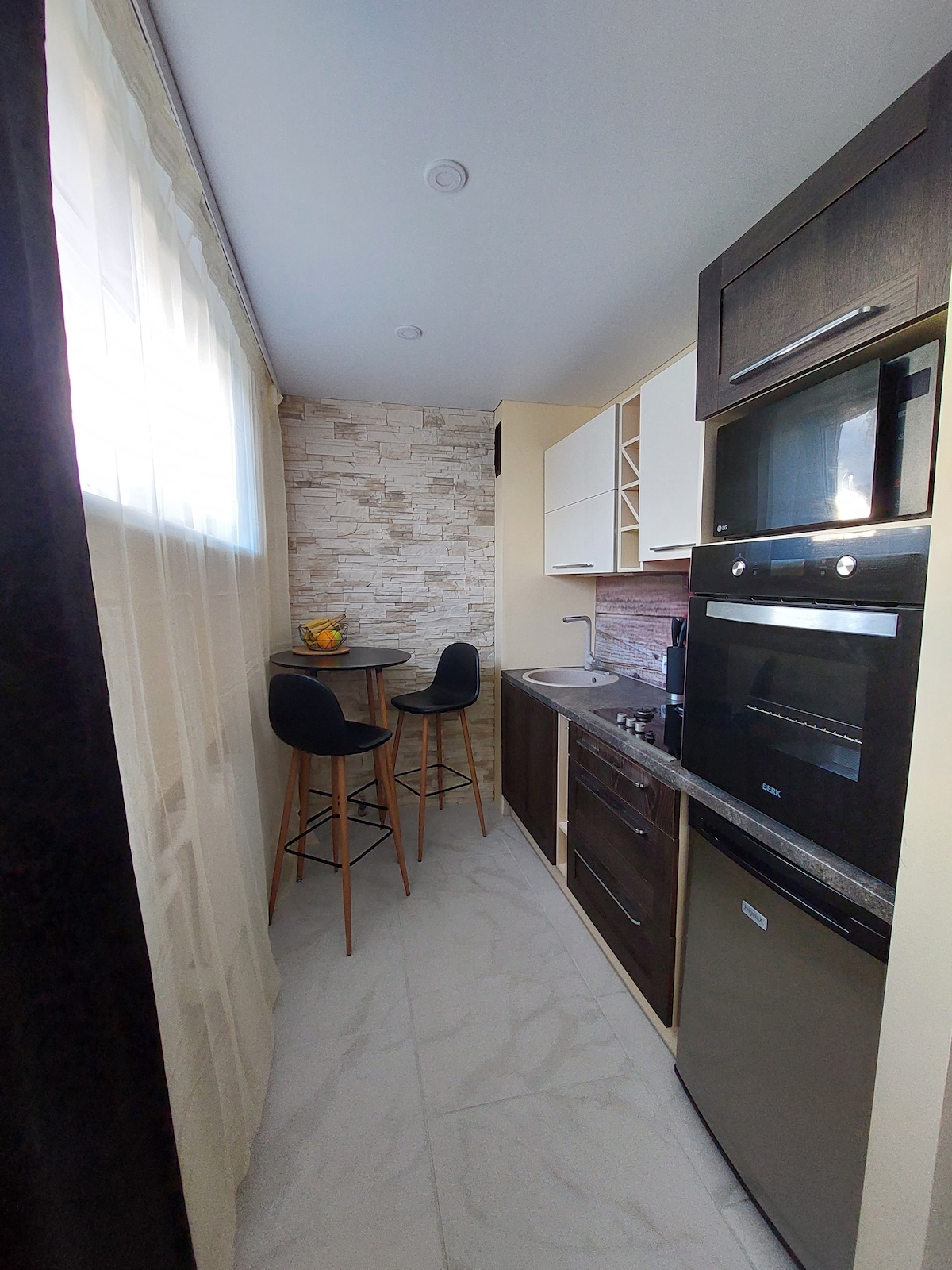 A compact kitchen features modern cabinetry and stainless steel appliances. A small round dining table with two tall chairs is positioned near a window, allowing for natural light. Light-colored stone walls provide a calming backdrop to the functional space.
