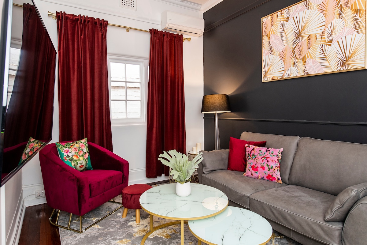 A stylish lounge area features a gray sofa adorned with vibrant pillows, alongside a round marble coffee table with gold accents. A striking burgundy armchair adds a pop of color, complemented by bold curtains. Natural light filters through a window, enhancing the room's contemporary design.