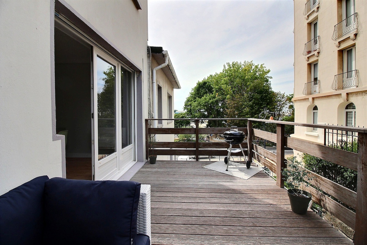 A spacious wooden terrace is visible, furnished with a comfortable seating area and a barbecue grill. Lush trees and greenery are seen in the background. Large glass doors open directly into the living area, creating a seamless connection to outdoor space.