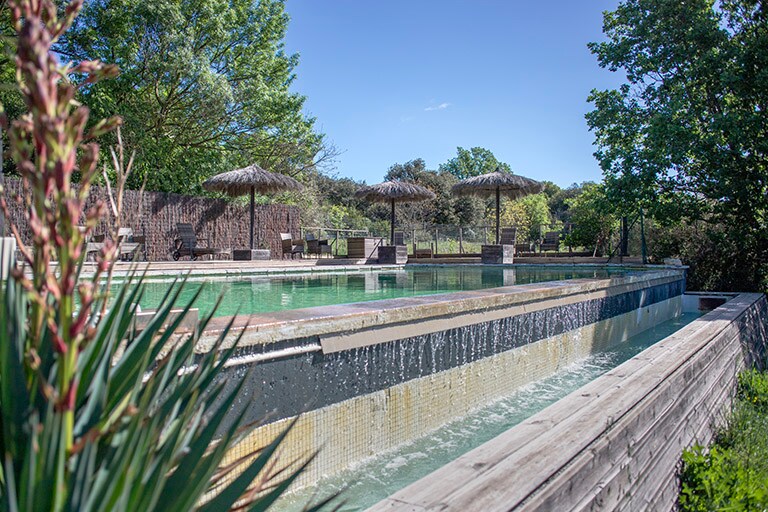 A spacious infinity pool is surrounded by wooden decking and shaded by parasols. Lush greenery and trees frame the area, creating a serene outdoor setting. Water cascades gently along the edges of the pool, enhancing the tranquil atmosphere.