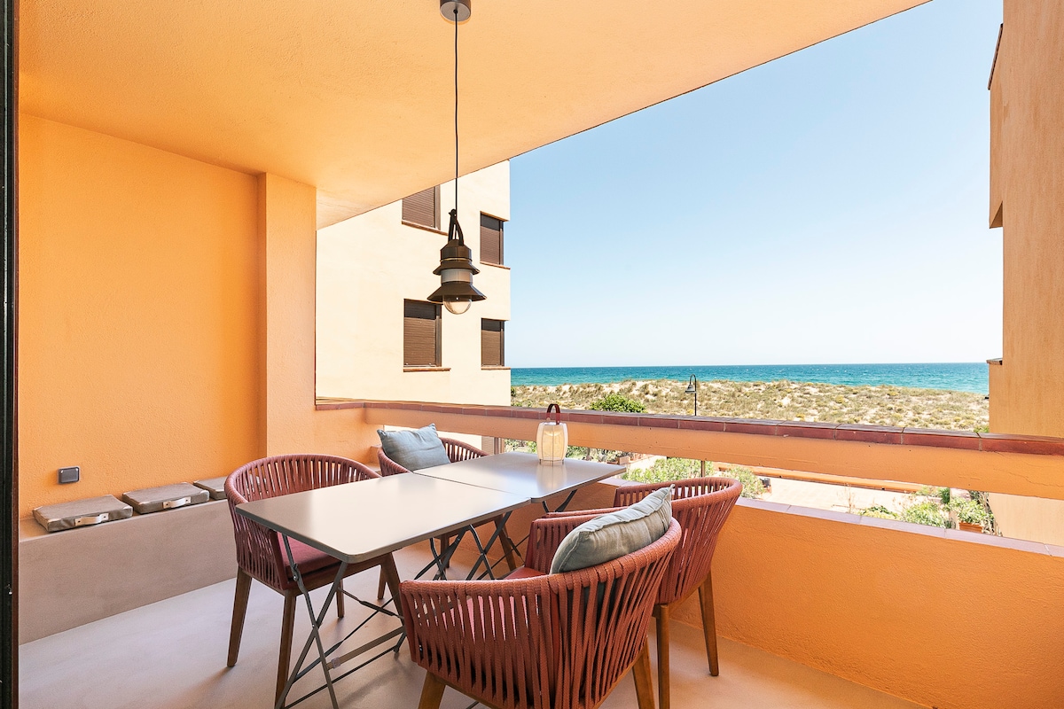A spacious terrace features a dining table accompanied by four comfortable chairs, overlooking scenic views of the beach and community area with lush greenery. Natural light floods the space through large windows, creating an inviting environment for relaxation.