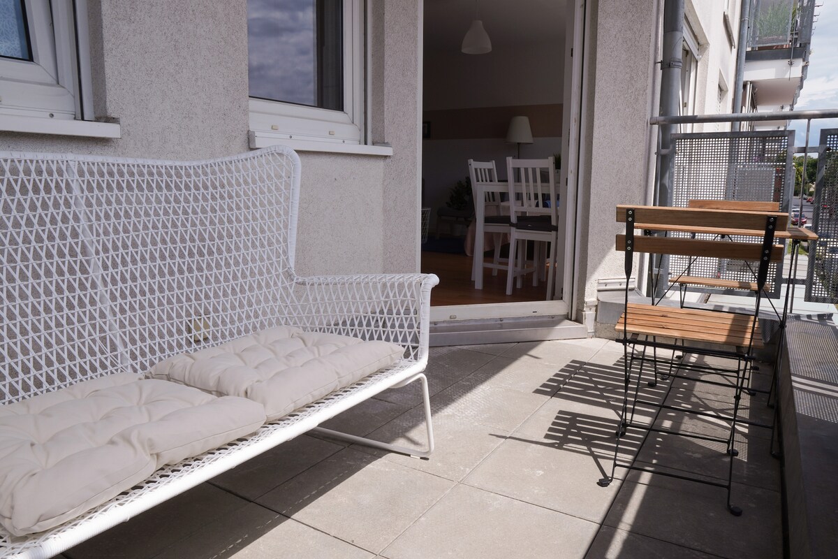 A small balcony features a white wicker bench with cushions alongside a compact outdoor table and two chairs. Sunlight illuminates the space, which offers a view of the surroundings, allowing for comfortable outdoor relaxation.