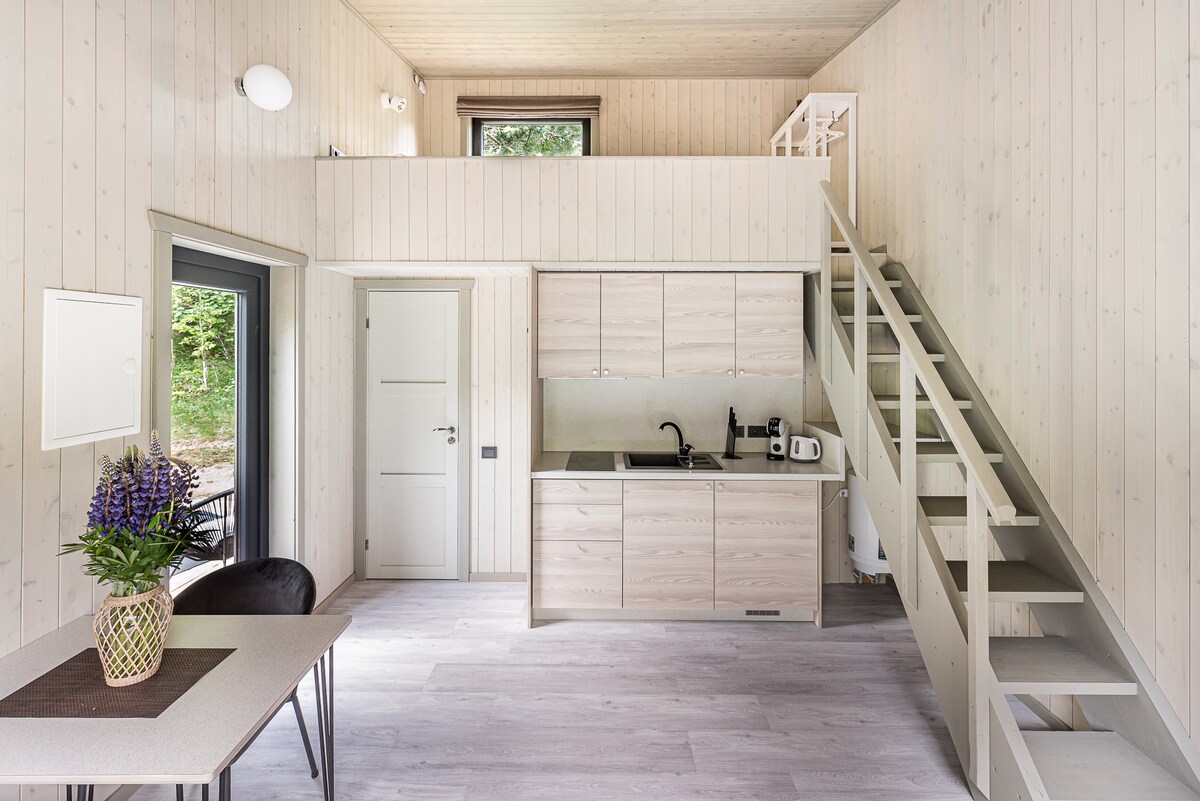 The interior space features light wood paneling and a functional design. A cozy dining table with a vase of flowers sits adjacent to a well-equipped kitchen, with essential appliances visible. A staircase leads to an upper level, adding a touch of height to the open area.