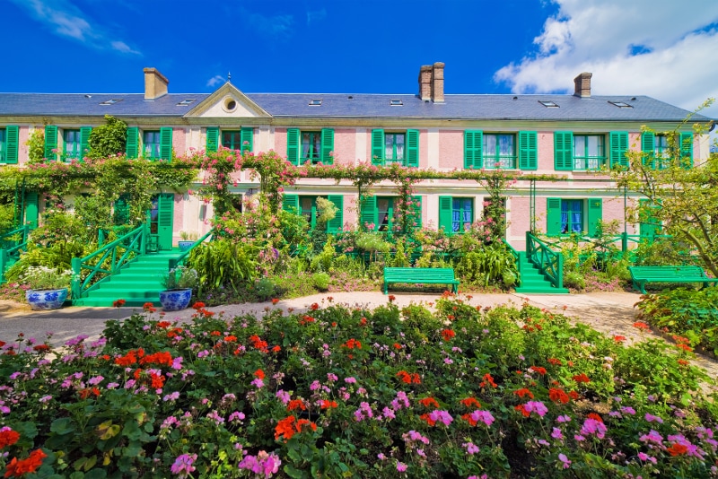 A colorful garden is presented in the foreground, featuring vibrant flowers and green foliage. Behind the garden, a two-story building with green shutters and doors is showcased, surrounded by climbing plants and trees under a bright blue sky.