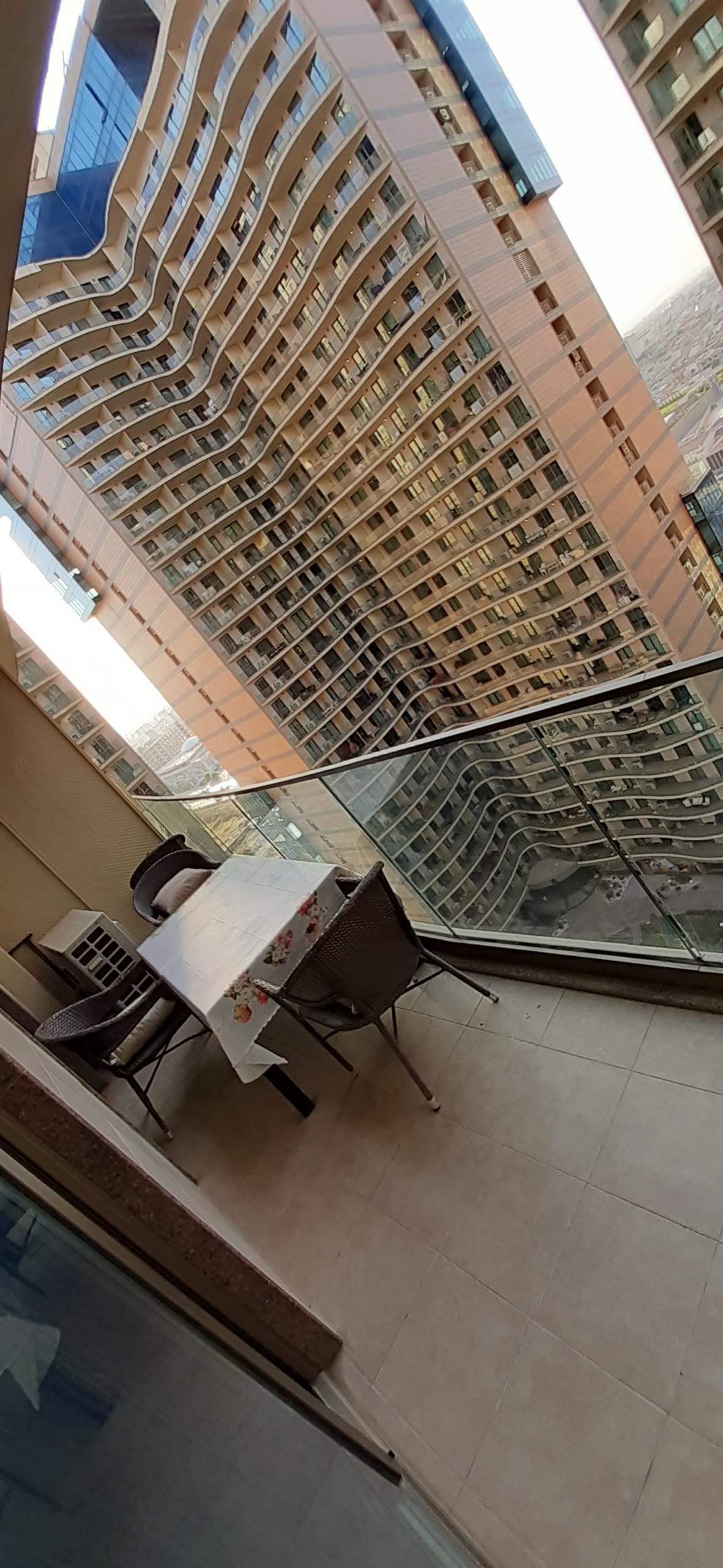 A balcony is shown with a glass railing, providing an expansive view of surrounding buildings. A small table is set with four chairs, and the tiled floor complements the urban setting, creating an inviting outdoor space.