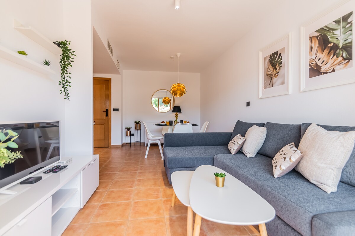 A modern living area features a comfortable grey sofa adorned with decorative pillows. A sleek coffee table sits in front, while a television is mounted on a white media unit. Natural light enters through a circular window, and potted plants add subtle greenery.