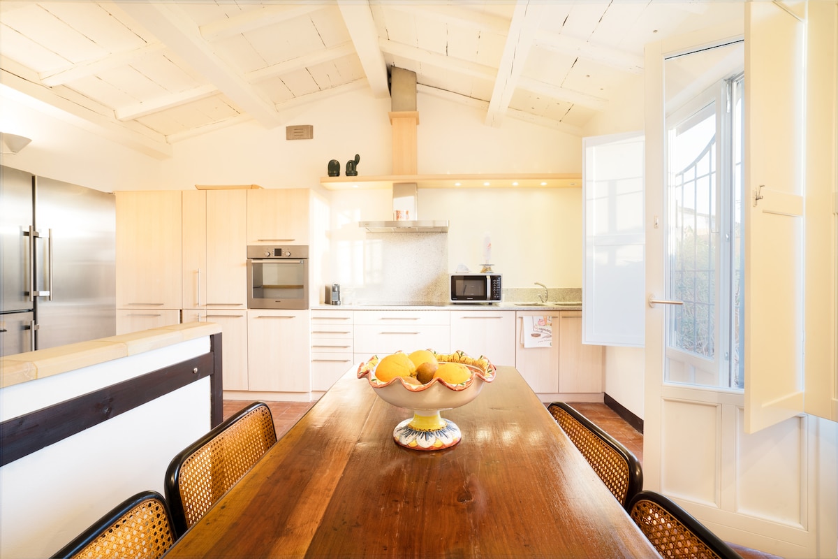 An airy kitchen is furnished with wooden cabinetry and modern appliances, including an oven and refrigerator. A spacious wooden dining table is centered with a fruit bowl. Natural light enters through large windows, highlighting the high ceiling with exposed beams.