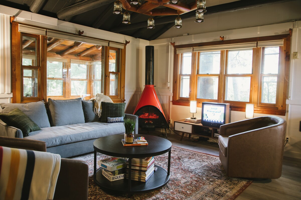 A comfortable living area is highlighted by a spacious sofa, complemented by an armchair. A round coffee table holds books and a small plant. A mid-century style electric fireplace adds warmth, while large windows illuminate the space and offer views of the outdoors.