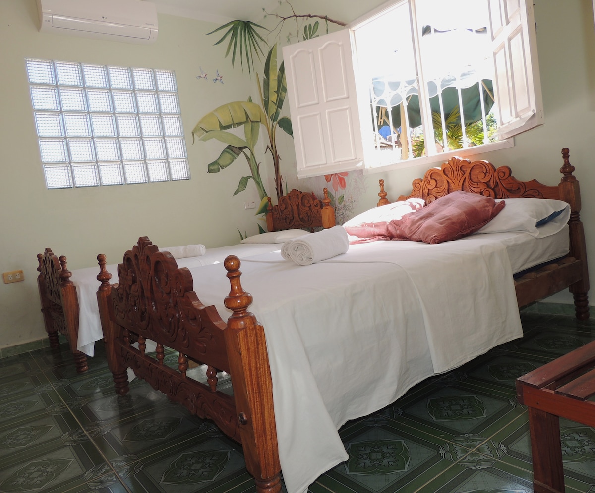 A well-lit room featuring two wooden beds, one double and one single, with light-colored bedding. Decorative wall art of tropical plants adds charm to the space. Windows allow natural light to fill the room, creating an inviting atmosphere with green tiled flooring.