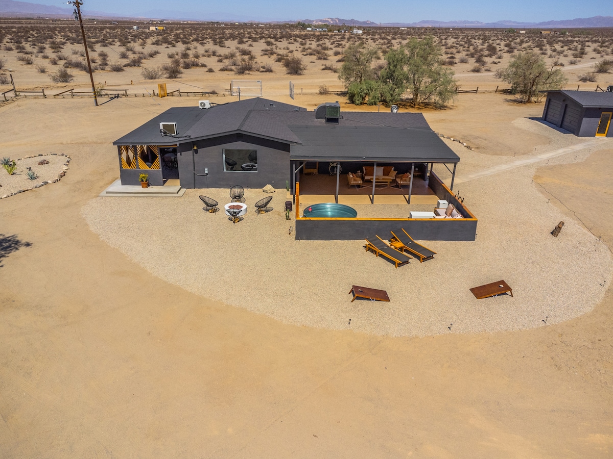 An aerial view of a contemporary dark-hued house surrounded by expansive, arid desert land. The property features a small pool, lounging chairs, and a fire pit area, all set against a backdrop of rugged terrain and distant mountains.