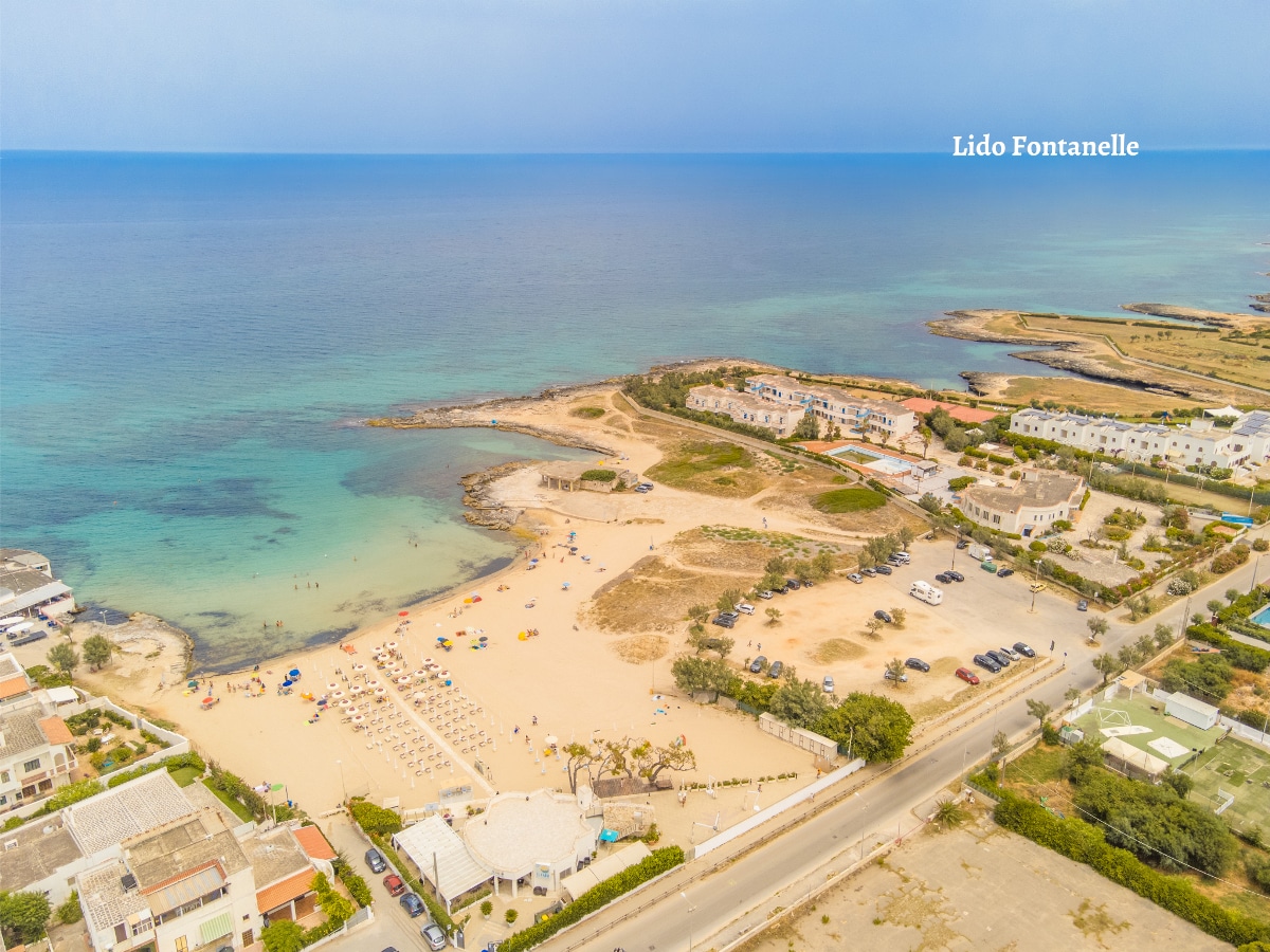 An aerial view of a sandy beach is presented, featuring a coastline lined with gentle waves. Sun loungers and umbrellas are arranged on the shore, with clear blue waters stretching into the distance. Nearby structures, including a parking area, are visible along the seaside.