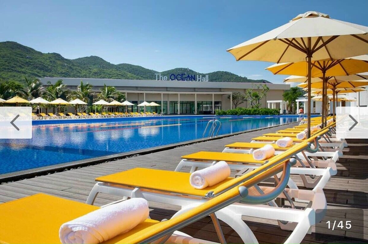 An expansive outdoor pool area is featured, lined with yellow sun loungers and white towels neatly rolled on each. The clear blue water reflects the sky, while a row of umbrellas provides shaded seating. Lush green hills are visible in the background.