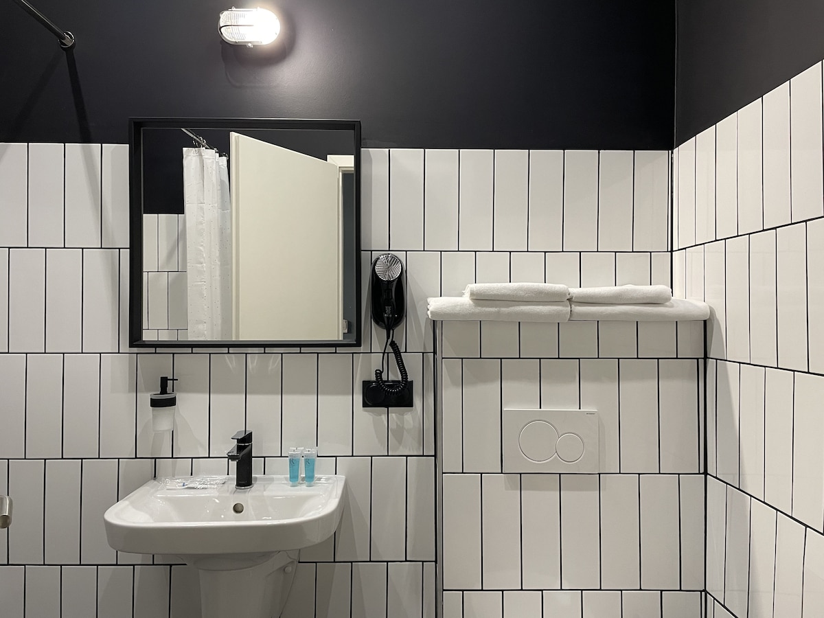 A compact bathroom features a white pedestal sink against a tiled wall with black grout. A large mirror reflects the space, and a shelf holds neatly arranged white towels. Personal care items, including two bottles, are placed on the sink. A hairdryer is mounted on the wall.