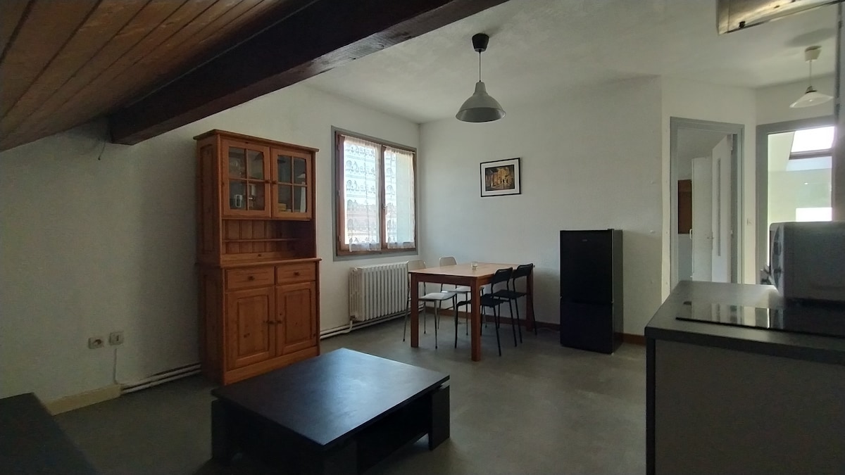 A spacious living area is shown, featuring a wooden cabinet against a wall and a dining table surrounded by chairs. A black refrigerator is positioned near a light-colored countertop. Natural light enters through a large window, illuminating the minimalist design with simple lines.