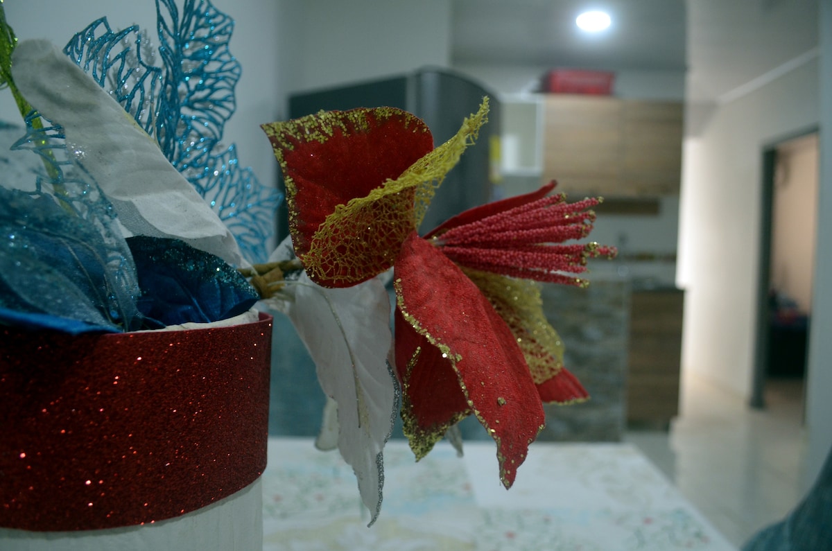 A decorative arrangement of colorful artificial leaves in red, blue, and gold is showcased in a white and red pot. In the background, a modern kitchen with wooden cabinets is partially visible, featuring dark countertops and appliances.