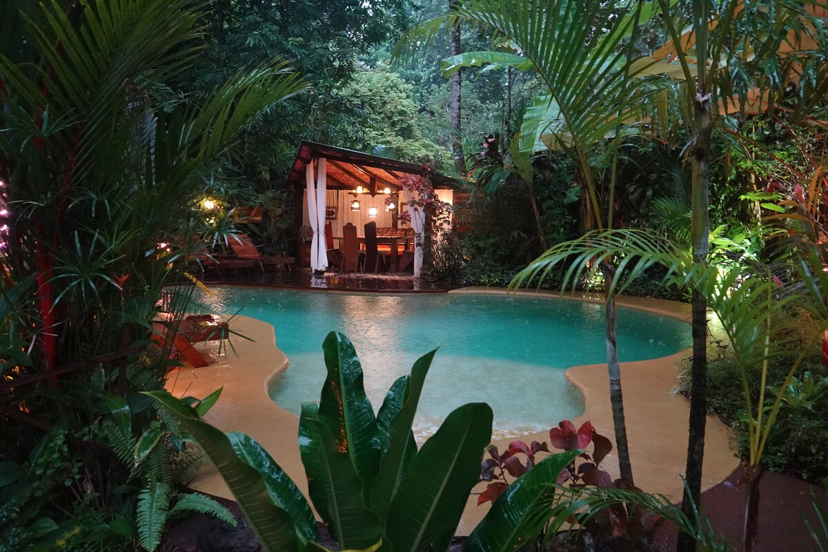 A serene pool area is surrounded by lush tropical foliage, featuring a covered dining area with seating. Soft lighting highlights the inviting water and natural setting, creating a tranquil atmosphere for relaxation and enjoyment.