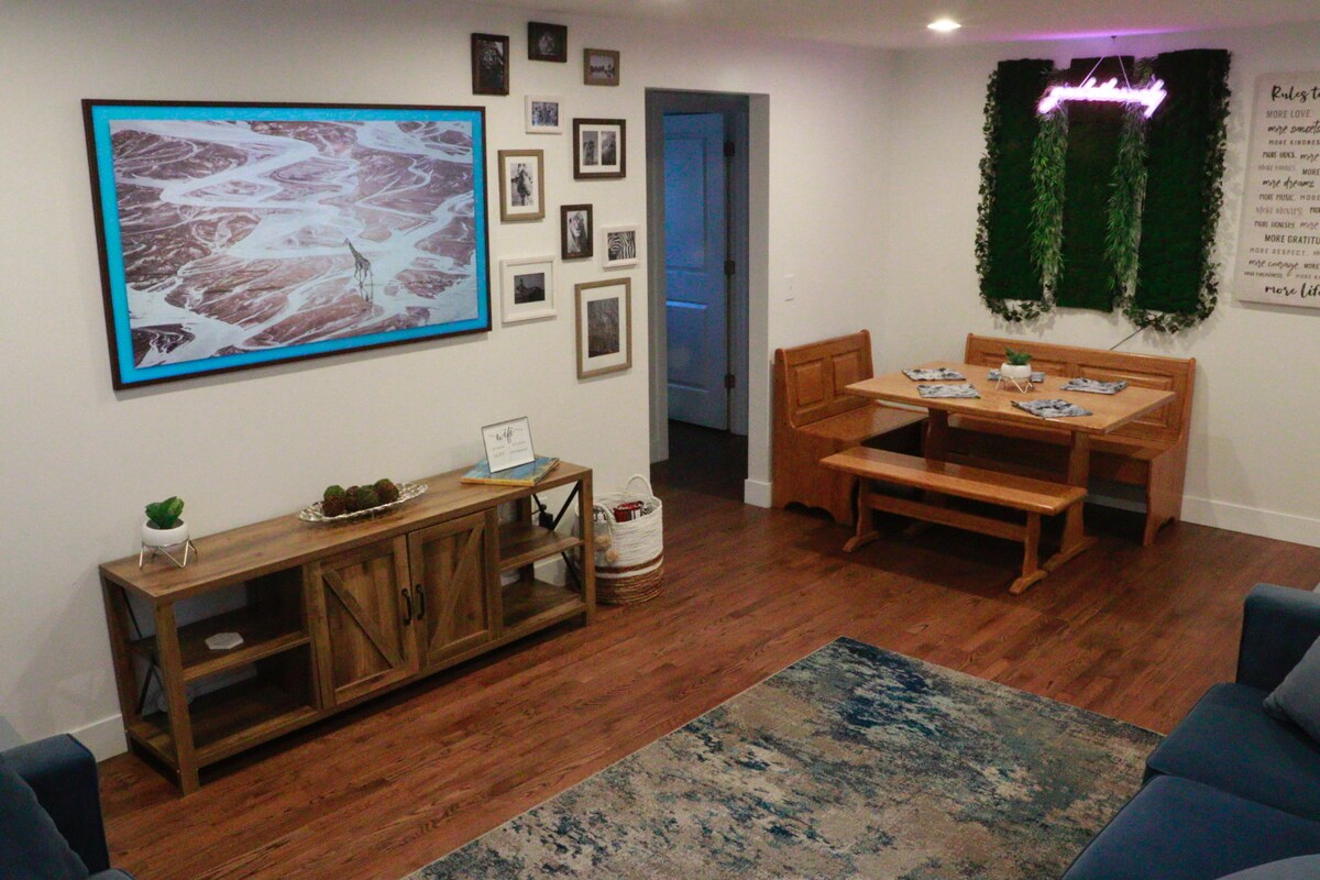 A main living area features a rustic wooden dining table with benches, accompanied by a console table displaying decorative items. A vibrant landscape artwork adorns the wall, alongside various framed photos. Soft seating is positioned to enhance relaxation, with warm lighting creating an inviting ambiance.