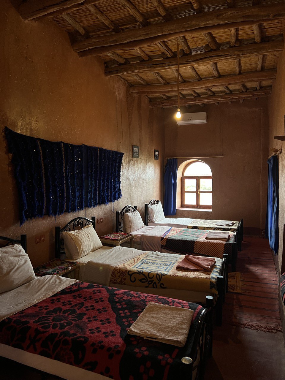 A private room is shown with four single beds arranged neatly. Traditional textiles decorate the beds, and a dark blue wall hanging adds texture to the earthy-toned walls. An arched window allows natural light to filter in, and a wooden beam ceiling enhances the rustic ambience.