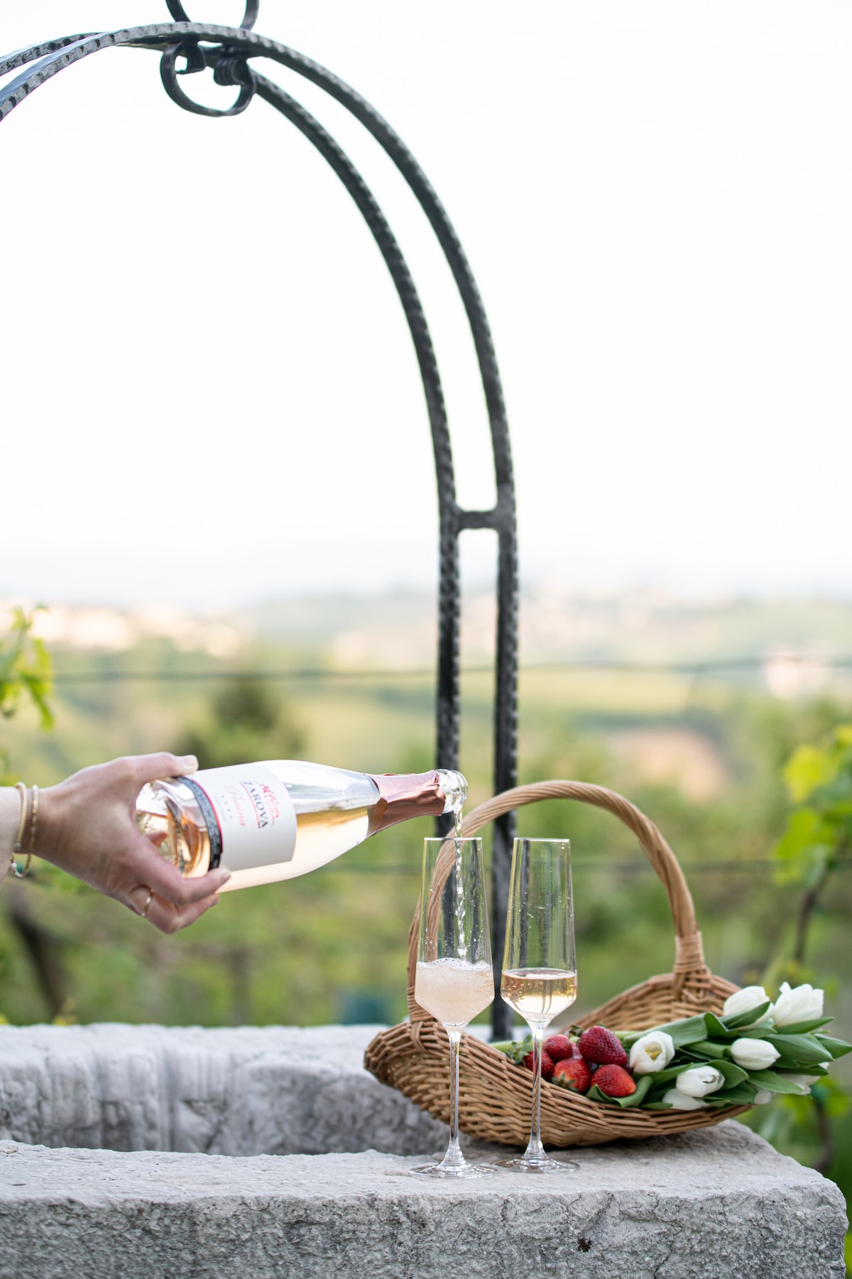 A bottle of sparkling rosé wine is being poured into elegant fluted glasses. Nearby, a wicker basket holds fresh strawberries and white tulips, set against a scenic backdrop of rolling hills and vineyards.