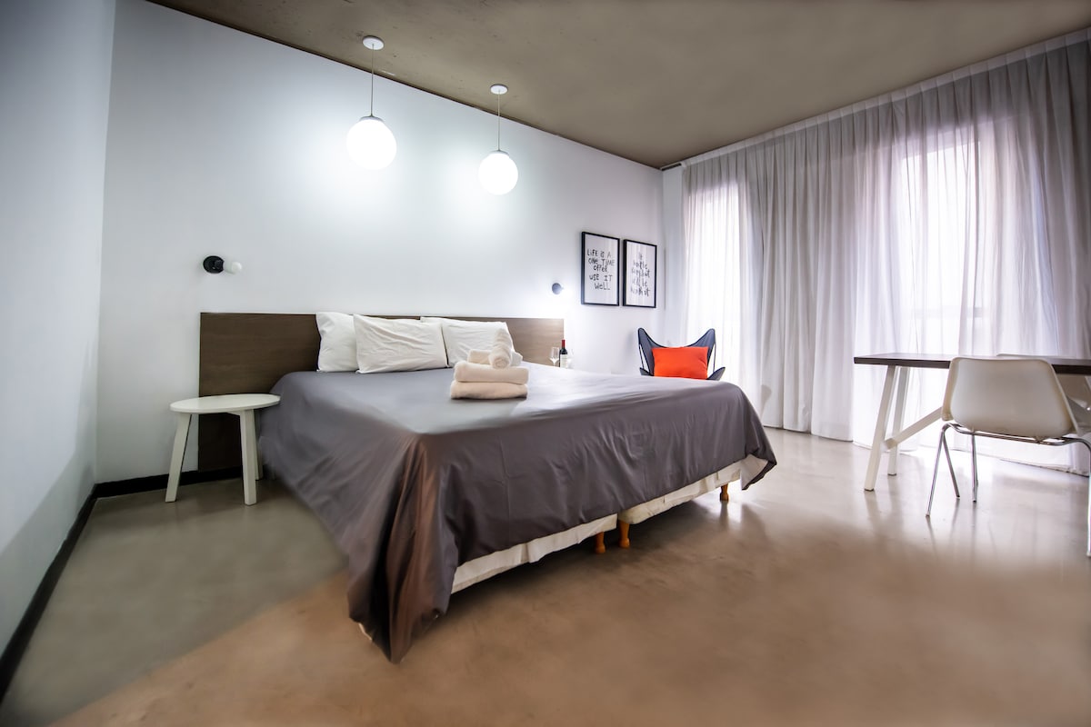 A spacious sleeping area features a large bed with neutral bedding, complemented by soft pendant lights overhead. A sheer curtain allows natural light to fill the room, while a minimalist chair and table provide a functional workspace.