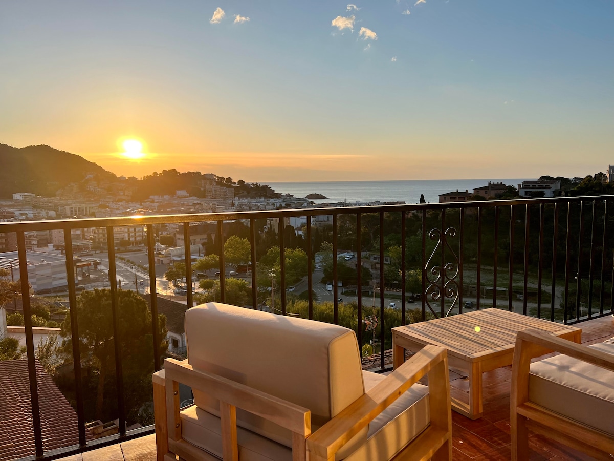 A private terrace is depicted, featuring two wooden chairs and a small table, overlooking a tranquil sea at sunrise. The horizon is painted with warm colors, while the town of Tossa is visible in the background, framed by lush greenery and rolling hills.