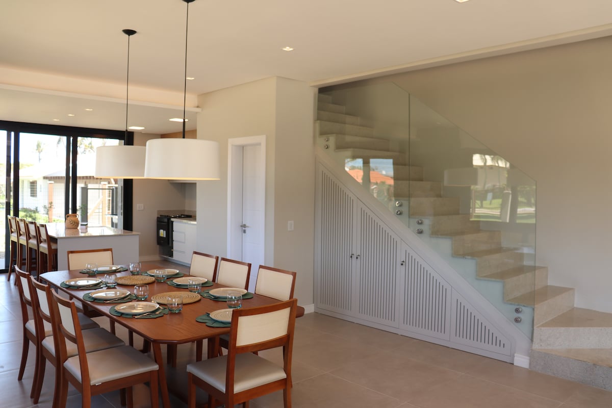 A spacious dining area features a large wooden table set for several guests, surrounded by light-toned chairs. A staircase with glass railing leads to the upper level, while large windows provide ample natural light and views of the outdoor space.