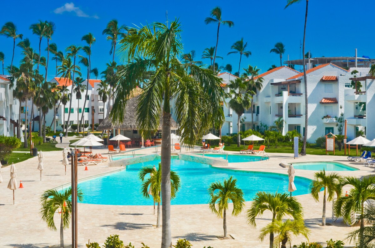 A spacious pool area is surrounded by palm trees and sun loungers under shaded umbrellas. The bright, clear blue sky enhances the inviting atmosphere of the well-maintained grounds, with white buildings visible in the background.
