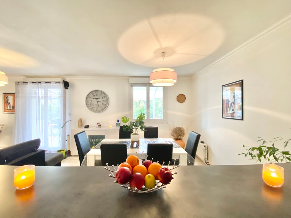 The dining area is illuminated by natural light streaming in from the nearby window. A round table with stylish black chairs is complemented by a decorative fruit bowl. Soft lighting from pendant lamps enhances the modern decor, and plants add a touch of greenery to the space.