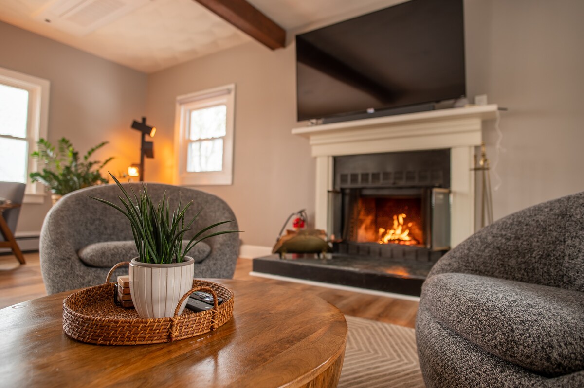 A cozy living room features two rounded armchairs positioned near a wooden coffee table. A simple potted plant rests on the table, while a fireplace creates a warm ambiance in the background. The room is brightened by natural light from nearby windows.