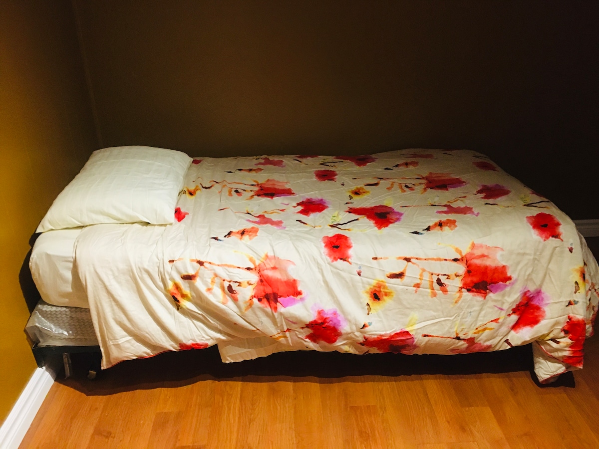 A spacious bed is dressed in a floral patterned comforter featuring shades of red and yellow. Soft pillows are neatly arranged at the head, complemented by warm walls and wooden flooring that contribute to a serene ambiance.