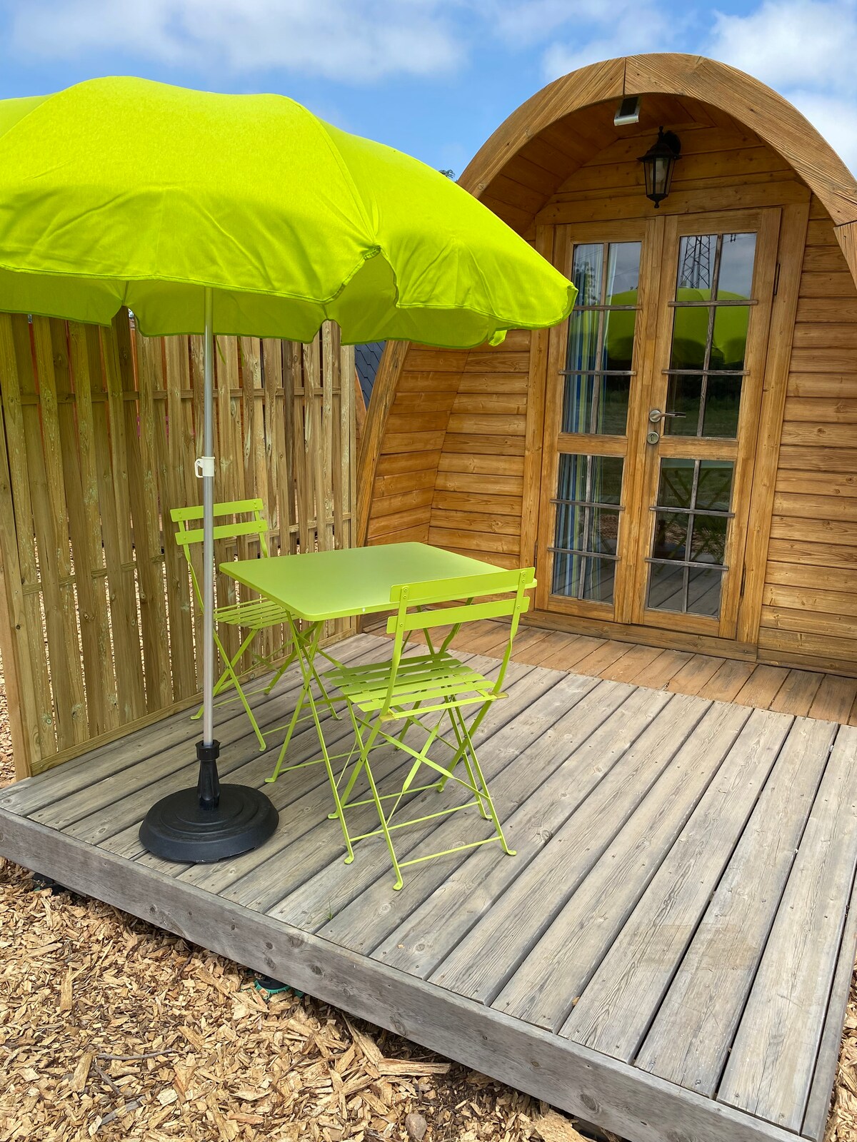 A wooden structure with a rounded roof is visible, accompanied by a small outdoor seating area. A bright green umbrella provides shade over a matching table and two chairs. The deck features wooden flooring surrounded by a natural wood fence.