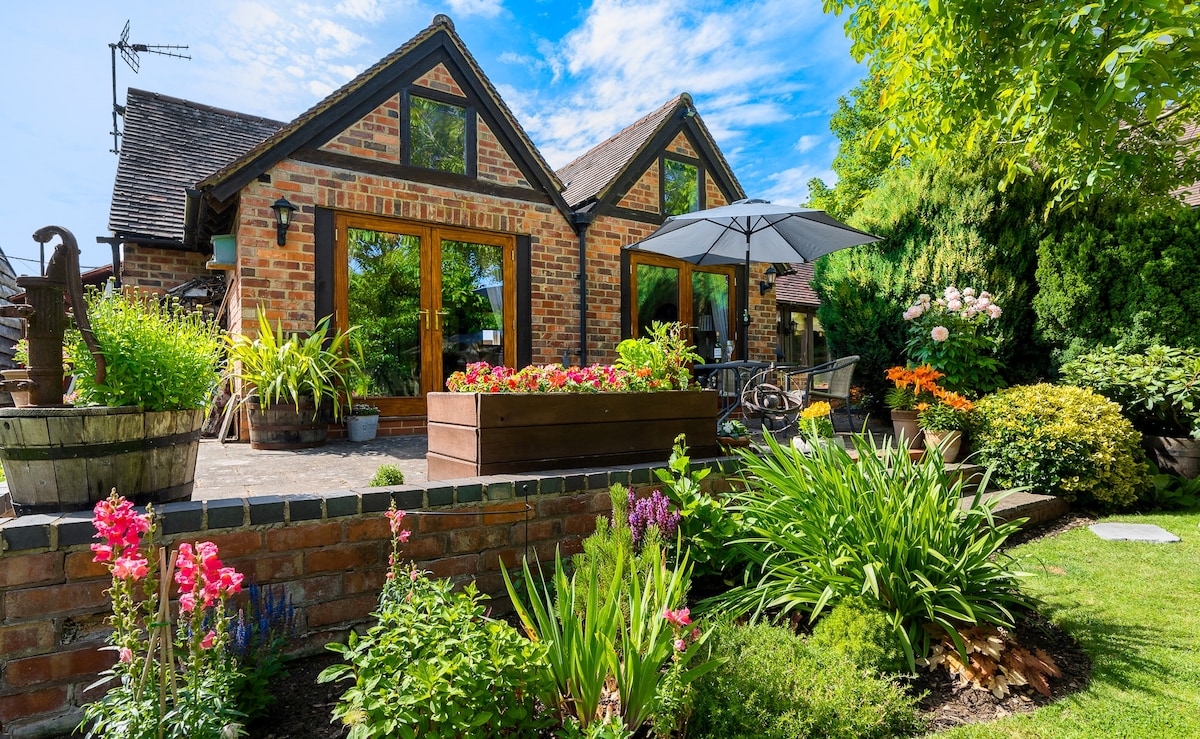 The exterior of a cottage-style guest suite features large windows framed by vibrant flowers and lush greenery. A shaded patio area with a table and chairs provides an inviting outdoor space, complemented by planters and a variety of colorful plants.