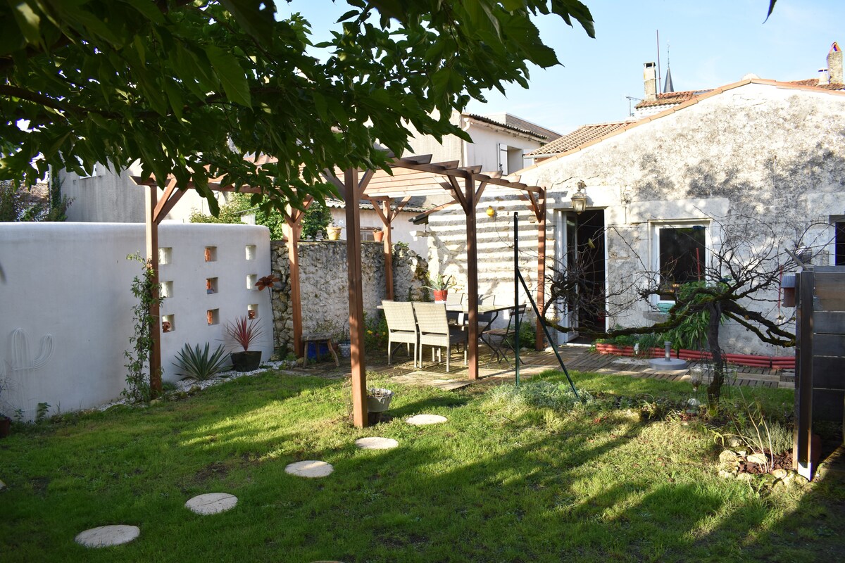 A tranquil garden space is visible, featuring a neatly manicured lawn bordered by various plants. A wooden pergola holds seating, providing an inviting area for relaxation. The rustic stone wall of the house is partially obscured by greenery, adding to the peaceful ambiance.