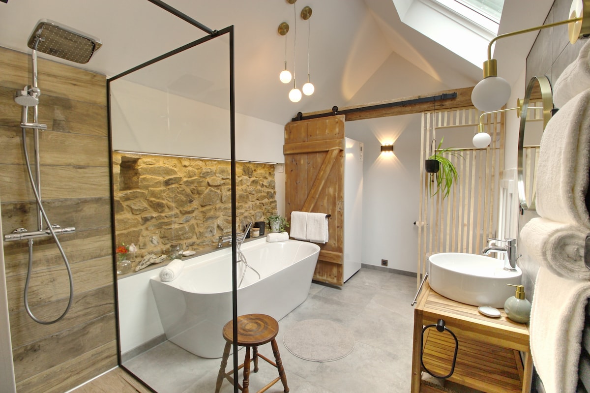 A spacious bathroom features a modern freestanding bathtub and a glass shower enclosure with sleek fittings. Natural stone walls add texture, complemented by wooden accents. Soft lighting from hanging fixtures and wall lamps creates a cozy ambiance. A stool is positioned beside the bathtub.