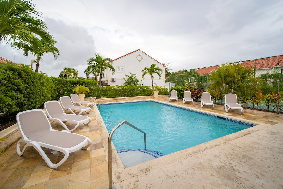 A shared swimming pool is surrounded by tropical landscaping, featuring multiple sun loungers positioned along the water's edge. The inviting pool is designed for relaxation, with steps leading into the clear water and a tranquil atmosphere created by the nearby greenery.