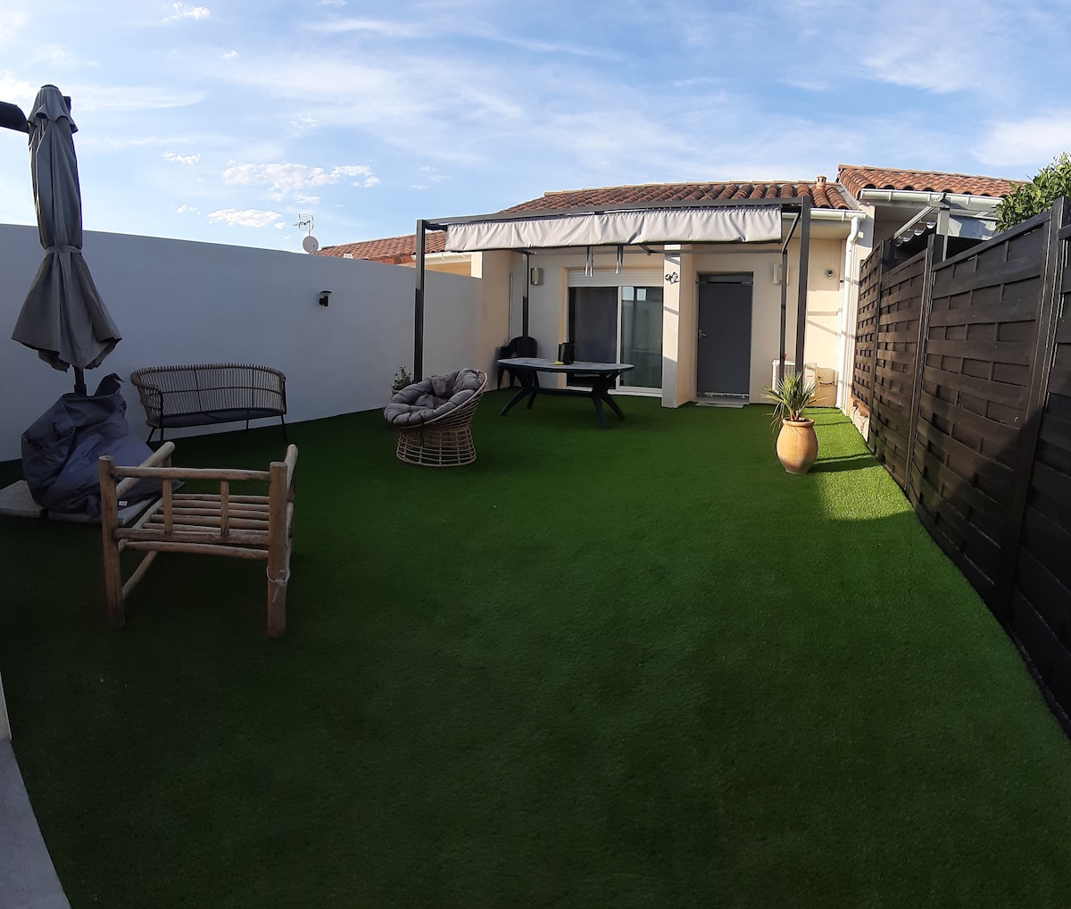 A spacious outdoor area is covered in artificial grass, featuring a comfortable seating arrangement with lounge chairs. A table is placed under a shaded area, while a parasol stands nearby. The backdrop consists of modern walls and a sliding door leading to a living space.