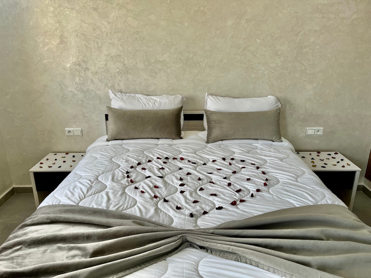 The bedroom features a double bed adorned with decorative rose petals, set against a textured wall. Two bedside tables flank the bed, each displaying decorative details. Soft, neutral-coloured pillows complement the bedding, while the overall setting offers a serene and gentle ambiance.