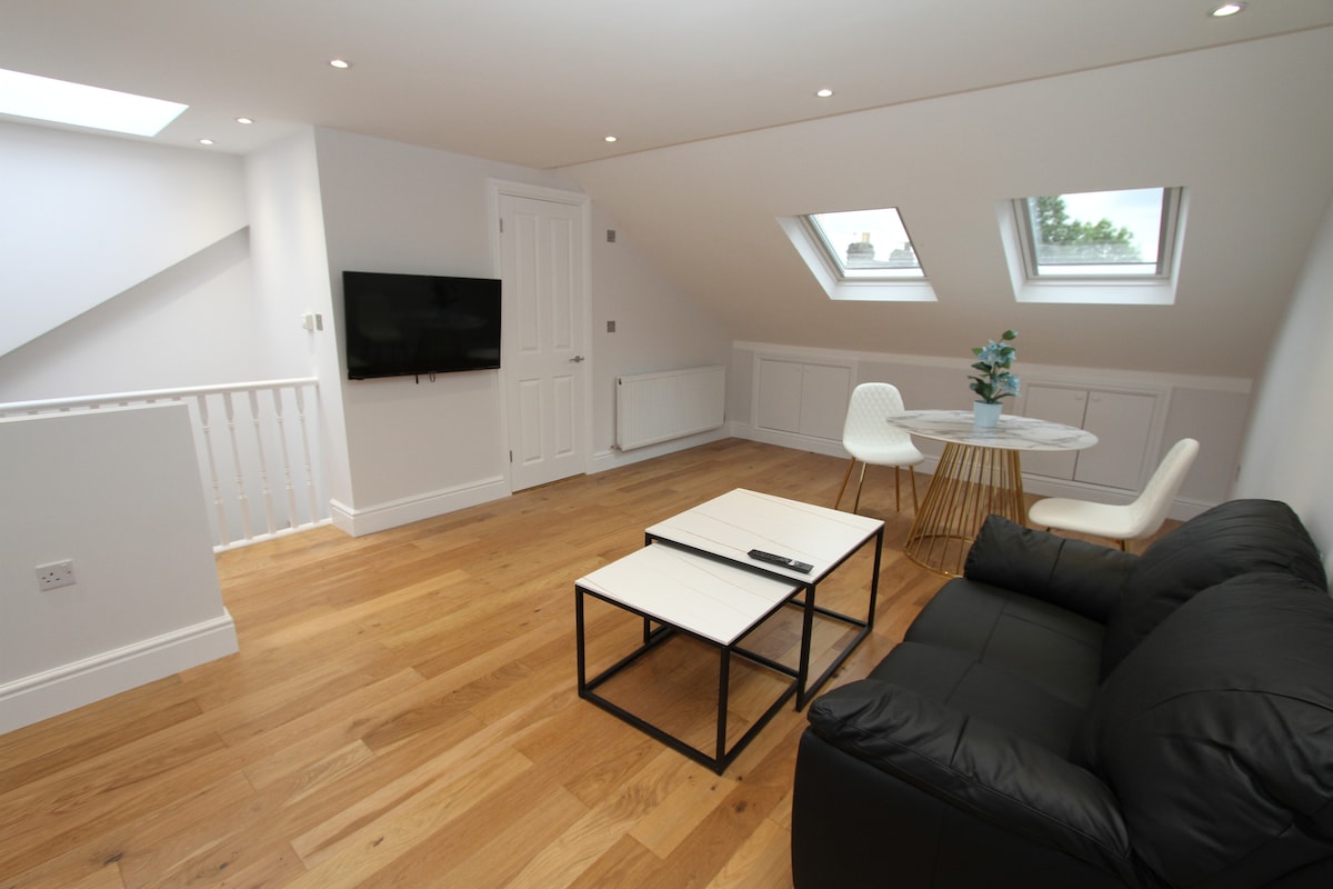 A spacious living area is illuminated by natural light from three skylights. A sleek black sofa sits across from a pair of modern coffee tables. A dining table for four is positioned nearby, complemented by white chairs and a decorative plant on the table.