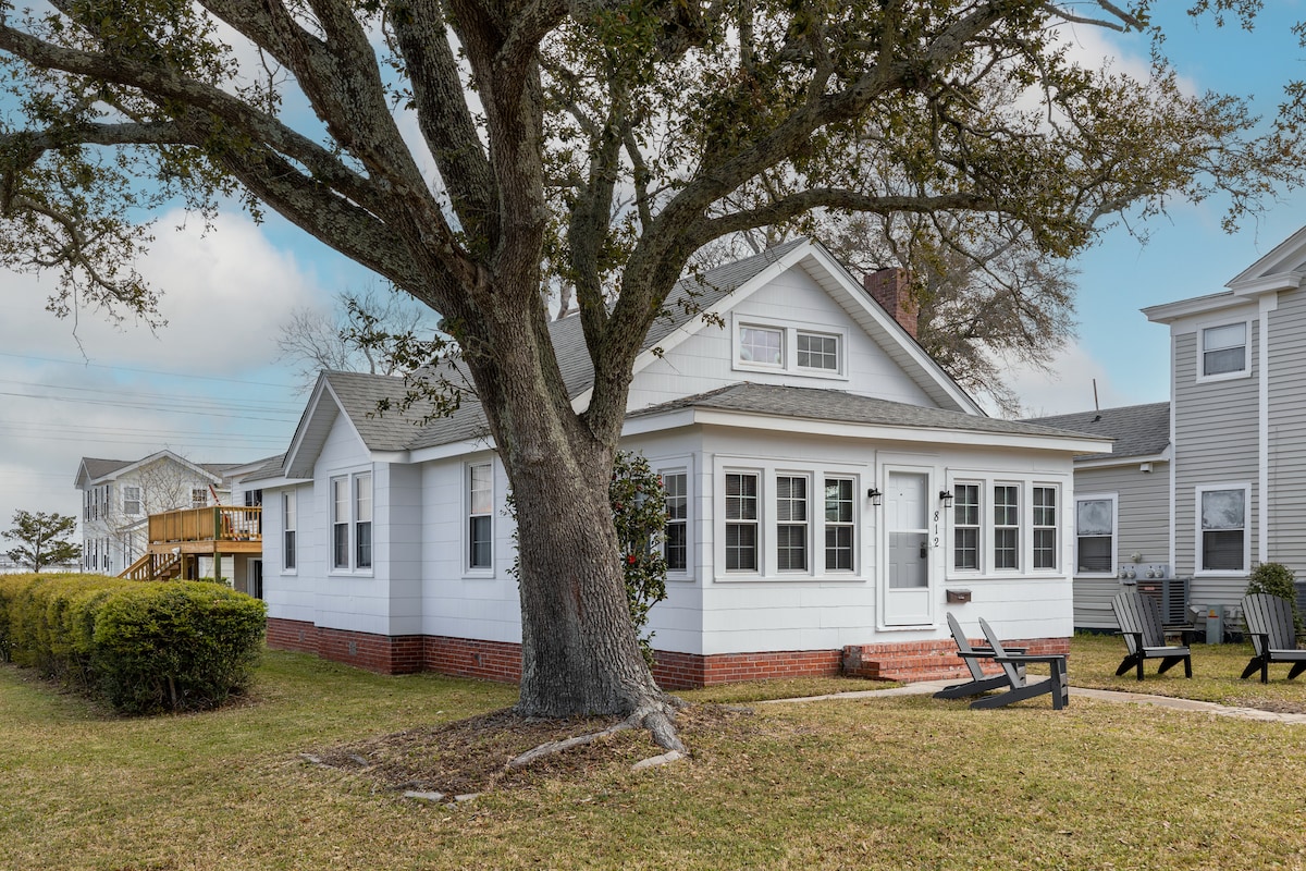 The charming cottage is nestled beneath a large tree, featuring a white exterior with multiple windows. A small porch with seating is visible at the entrance, while a well-kept lawn surrounds the home. Neighboring homes are seen in the background.