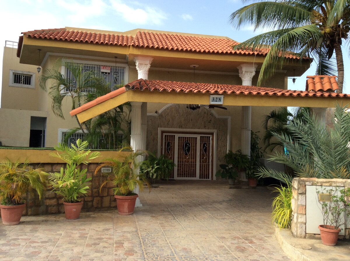 The exterior of a spacious residential villa is visible, featuring a terracotta roof and decorative columns. Planters with tropical plants line the entrance, leading to double doors adorned with glass motifs. Sunlight filters through, highlighting the welcoming ambiance of the property.