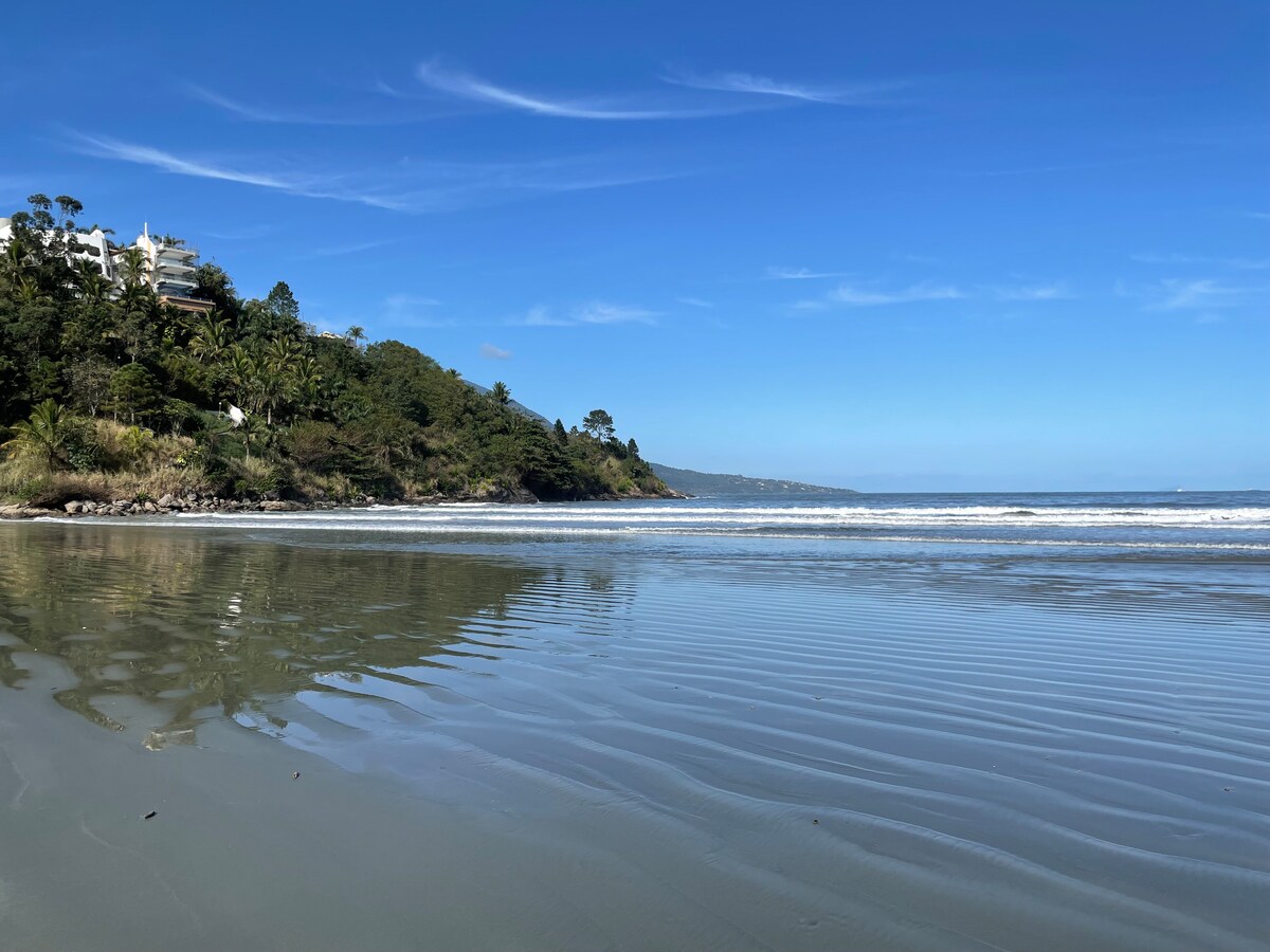 A tranquil beach scene features gentle waves lapping along a soft sandy shore under a clear blue sky. Lush greenery and hills can be seen in the background, reflecting a serene atmosphere, ideal for relaxation and leisure activities.