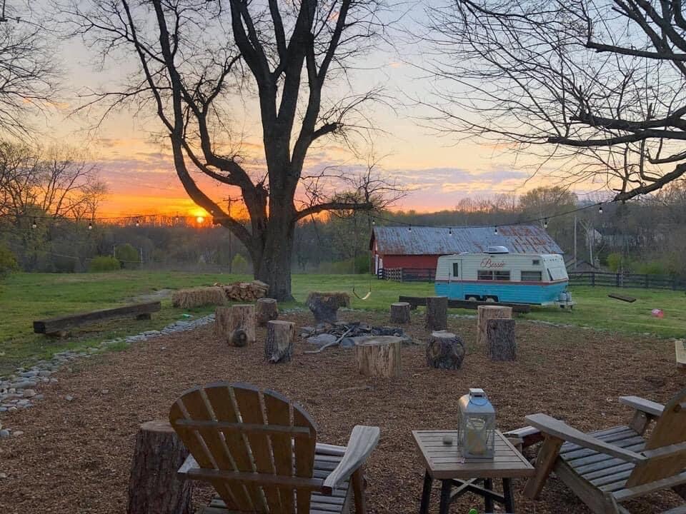 An outdoor seating area features two wooden chairs facing a fire pit surrounded by log stools. The scene is set against a vibrant sunset, with an old-fashioned trailer and a red barn visible in the background. Trees frame the tranquil landscape.